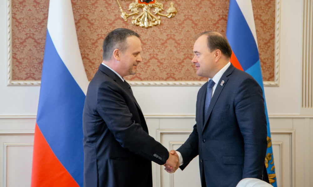 Двое мужчин в деловых костюмах пожимают руки на фоне российских флагов