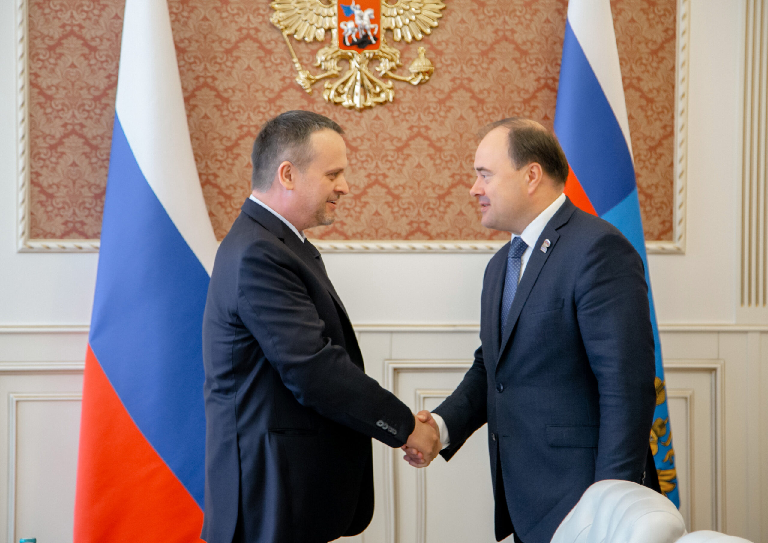 Двое мужчин в деловых костюмах пожимают руки на фоне российских флагов