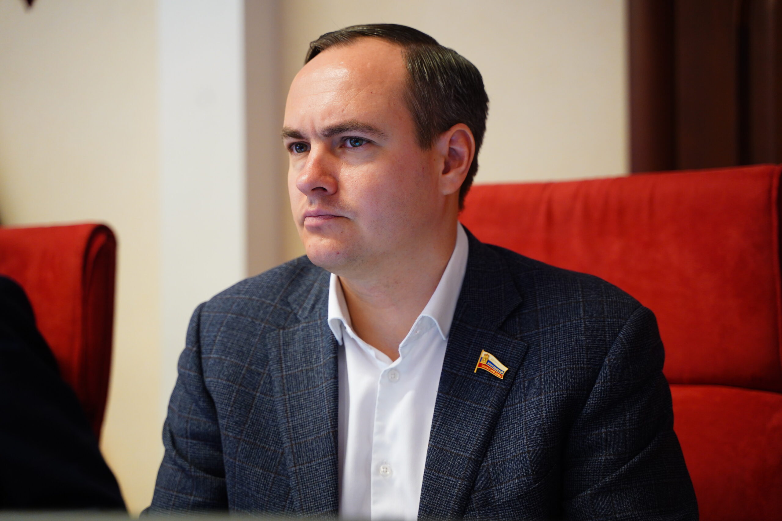 Мужчина в пиджаке с депутатским значком сидит в красном кресле