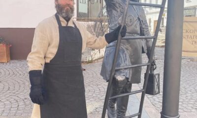 Мужчина в фартуке стоит у скульптуры фонарщика с лестницей в старом центре Рыбинска