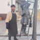 Мужчина в фартуке стоит у скульптуры фонарщика с лестницей в старом центре Рыбинска