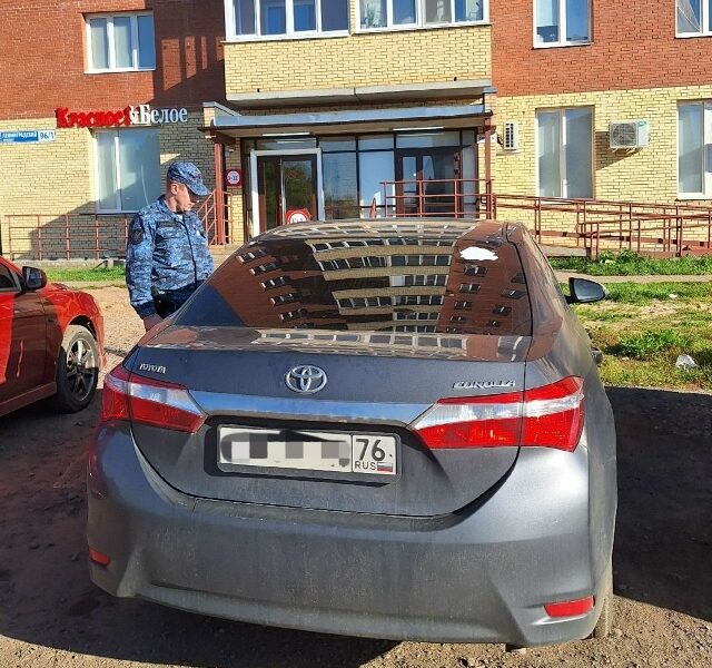 Серая Toyota Corolla у жилого дома, рядом стоит сотрудник в форме
