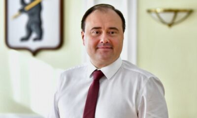 Артем Молчанов в белой рубашке и бордовом галстуке на фоне герба Ярославля