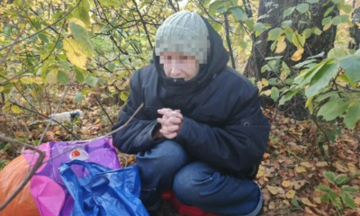 Пожилая женщина в куртке и шапке сидит в осеннем лесу рядом с сумками