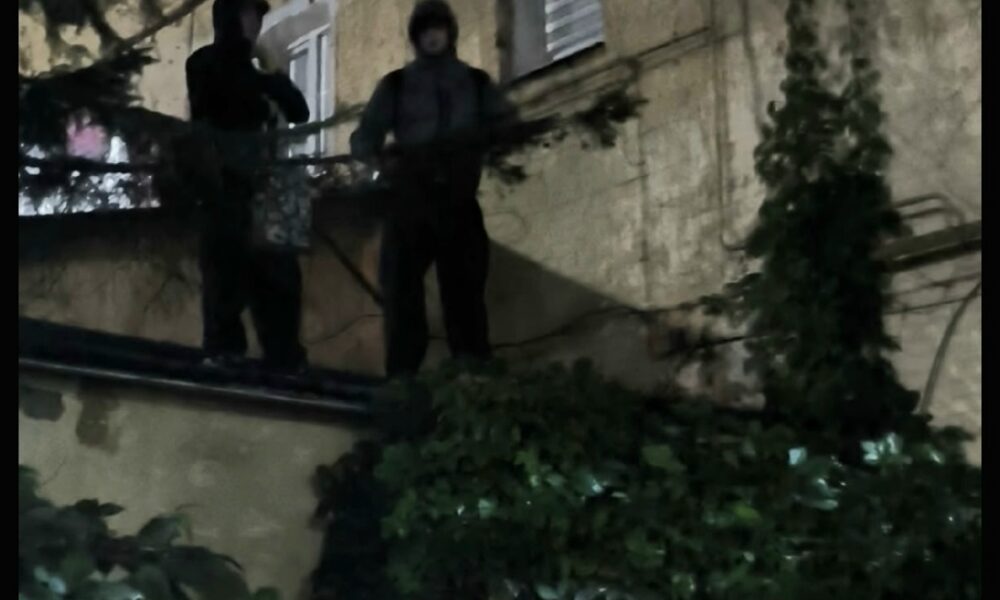 Двое людей в капюшонах стоят ночью на выступе у стены жилого дома