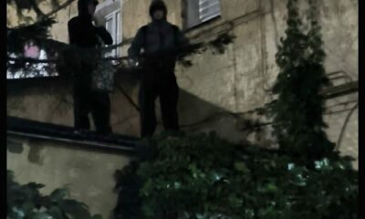 Двое людей в капюшонах стоят ночью на выступе у стены жилого дома