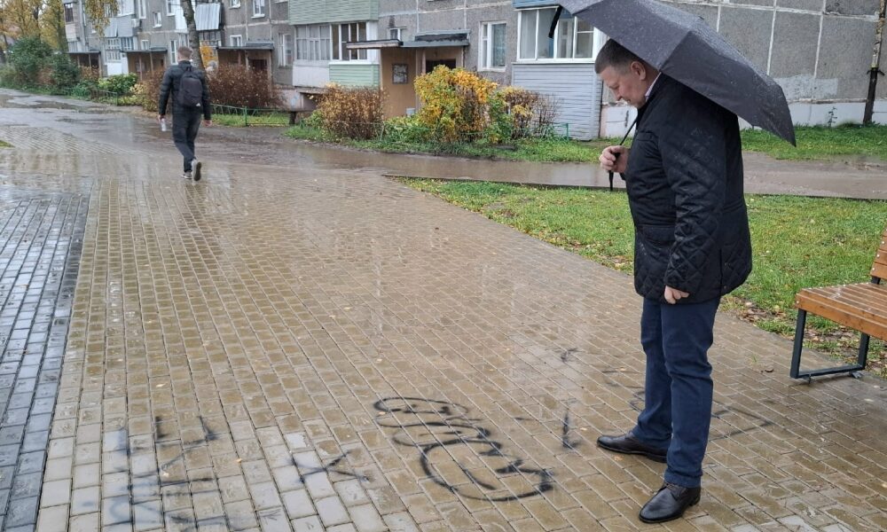 Мужчина с зонтом осматривает надписи вандалов на тротуаре у жилого дома