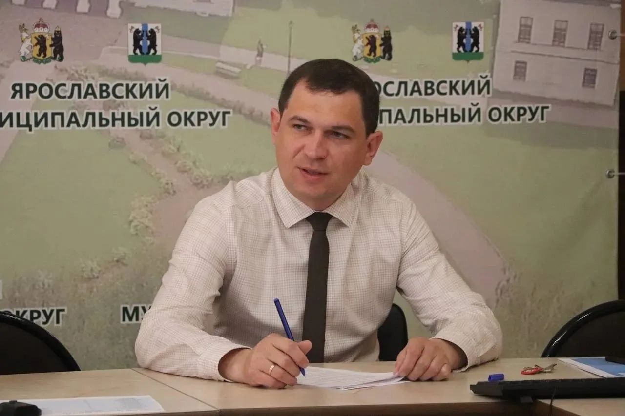Мужчина в рубашке и галстуке подписывает документы на фоне баннера Ярославского муниципального округа