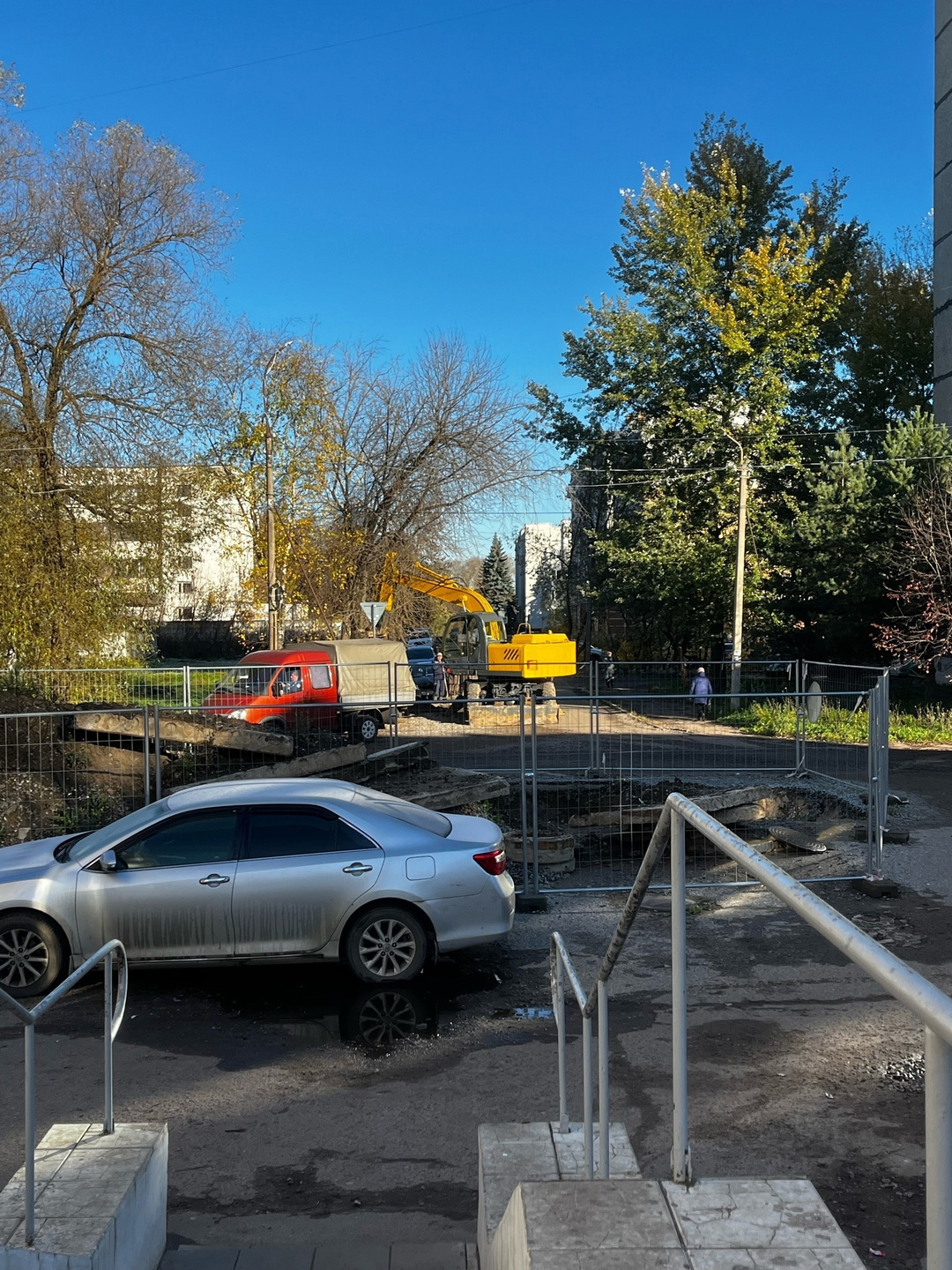 Экскаватор на огороженной строительной площадке во дворе среди автомобилей и осенних деревьев