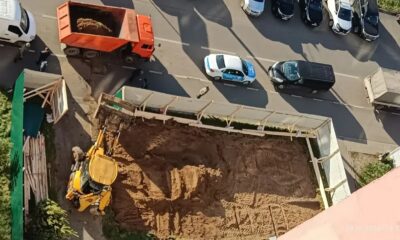 Экскаватор и самосвал на огороженной стройке рядом с жилым домом и припаркованными машинами